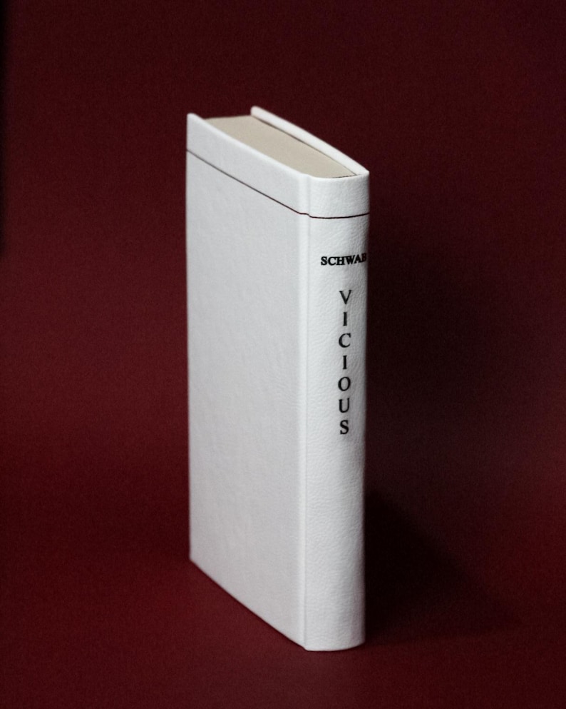 May include: A hardcover book with a white cover and the title "VICIOUS" printed vertically in black. The author's name, "SCHWAB", is printed above the title. The book is set against a dark red background.
