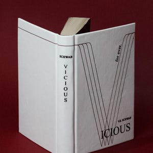 May include: A hardcover book titled "Vicious" by V.E. Schwab, with a white cover and black text. The book is open, revealing the pages. The cover features a stylized "V" design and the words "for ever."