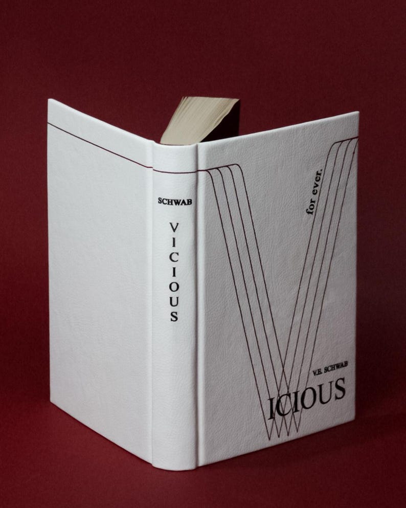 May include: A hardcover book titled "Vicious" by V.E. Schwab, with a white cover and black text. The book is open, revealing the pages. The cover features a stylized "V" design and the words "for ever."