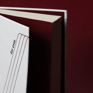 May include: Close-up of a book with white pages and a burgundy cover. The text "for ever." is printed vertically on the left side of the open book. The pages are cream-colored, and the background is a deep burgundy.