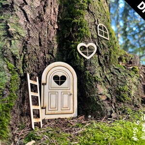 May include: DIY kit for a fairy door, featuring a wooden door with a heart-shaped window, a ladder, and a heart-shaped window. The kit is displayed against a tree trunk with moss.