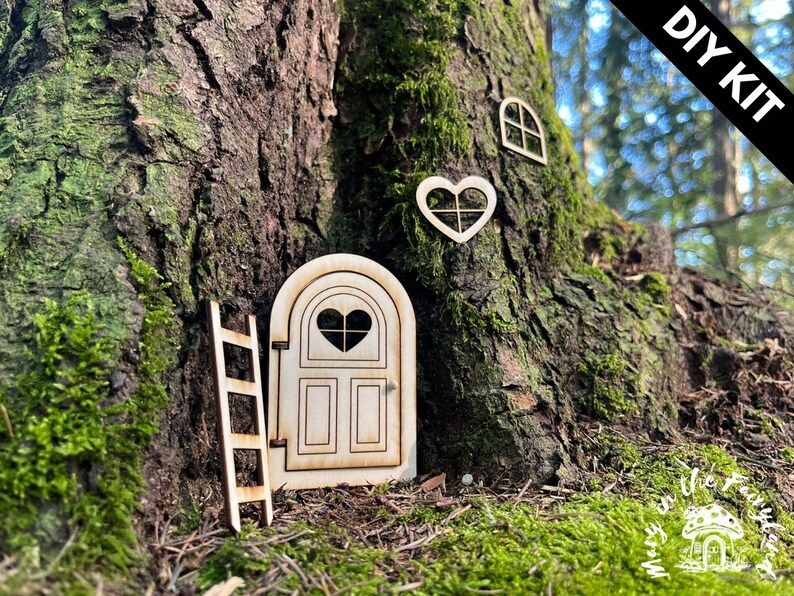 May include: DIY kit for a fairy door, featuring a wooden door with a heart-shaped window, a ladder, and a heart-shaped window. The kit is displayed against a tree trunk with moss.