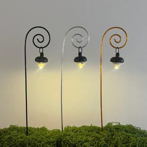 May include: Three miniature garden lights with a black, silver, and gold finish. Each light has a spiral top and a small, black lamp with a glowing yellow light. The lights are set in a bed of green moss.