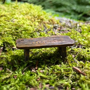 May include: A miniature wooden table with a rustic brown finish and a wood grain pattern. The table has three legs and is sitting on a bed of green moss.