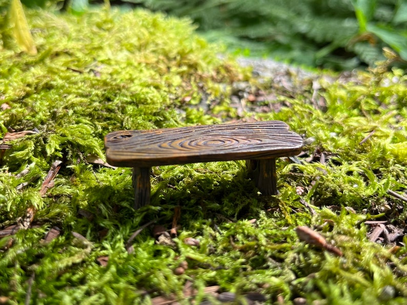 May include: A miniature wooden table with a rustic brown finish and a wood grain pattern. The table has three legs and is sitting on a bed of green moss.