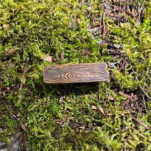 May include: A small wooden plaque with a wood grain pattern. The plaque is brown with a light brown wood grain pattern. The plaque is sitting on a bed of green moss.
