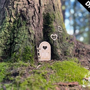 May include: DIY kit for a fairy door, featuring a wooden door with a heart-shaped window, a ladder, and a heart-shaped window. The door is set into the base of a tree trunk.