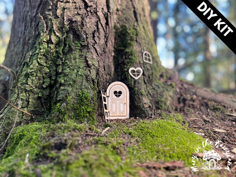 May include: DIY kit for a fairy door, featuring a wooden door with a heart-shaped window, a ladder, and a heart-shaped window. The door is set into the base of a tree trunk.
