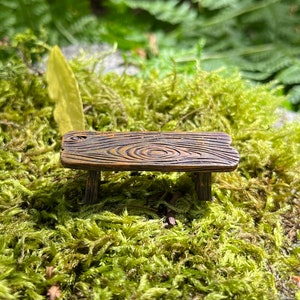 May include: A miniature wooden bench with a rustic brown finish and a natural wood grain pattern. The bench is sitting on a bed of green moss.
