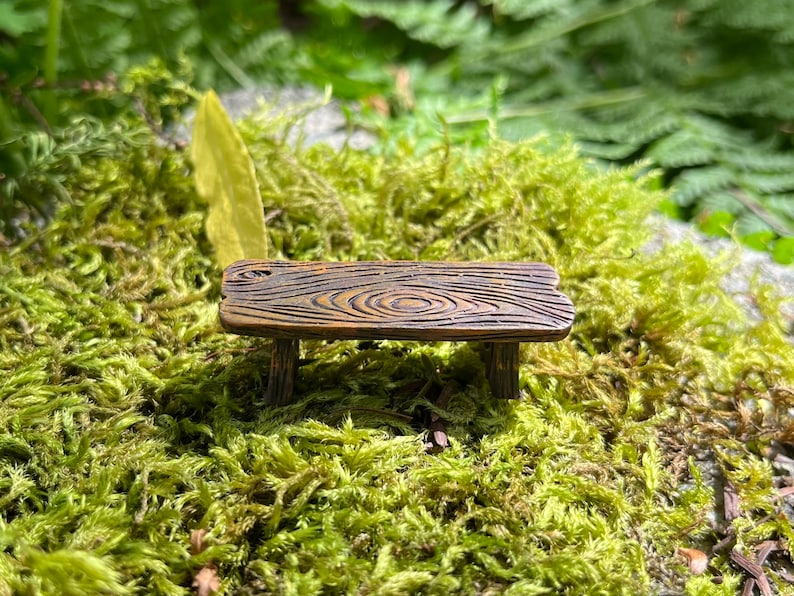 May include: A miniature wooden bench with a rustic brown finish and a natural wood grain pattern. The bench is sitting on a bed of green moss.