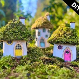May include: Three whimsical fairy houses with moss-covered roofs and colorful doors. The houses are painted light blue with yellow, pink, and blue doors. The image includes a sign and the text "DIY KIT". The houses are surrounded by green moss.
