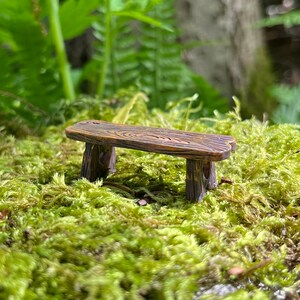 May include: A miniature wooden bench with a rustic brown finish, sitting on a bed of green moss.