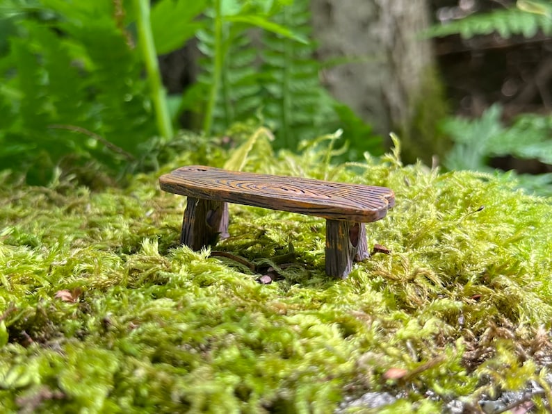 May include: A miniature wooden bench with a rustic brown finish, sitting on a bed of green moss.