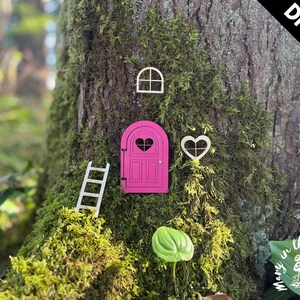 May include: DIY kit for a fairy door, featuring a pink door with a heart-shaped window, a ladder, a window, and a heart-shaped window. The kit is displayed on a tree trunk covered in moss.