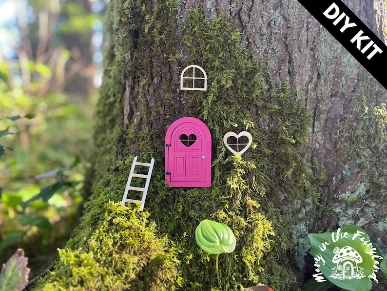 May include: DIY kit for a fairy door, featuring a pink door with a heart-shaped window, a ladder, a window, and a heart-shaped window. The kit is displayed on a tree trunk covered in moss.