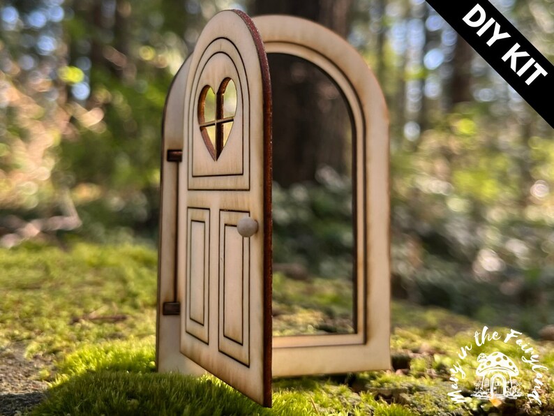 May include: DIY kit for a wooden fairy door with a heart-shaped window and a knob. The door is open and has a light wood finish.