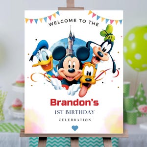 May include: A rectangular birthday sign featuring Mickey Mouse, Donald Duck, Goofy, and Pluto in front of a castle. The text reads WELCOME TO THE, Brandon's, 1ST BIRTHDAY, CELEBRATION, with colorful bunting at the top and a small blue heart at the bottom.