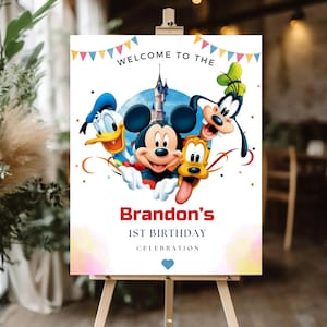 May include: A rectangular sign on a wooden easel featuring colorful bunting at the top and the text WELCOME TO THE. Below, illustrations of Mickey Mouse, Donald Duck, Goofy, and Pluto appear in front of a castle. Text reads Brandon's 1ST BIRTHDAY CELEBRATION.