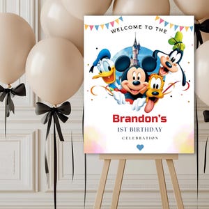 May include: A rectangular birthday sign on a wooden easel featuring Mickey Mouse, Donald Duck, Goofy, and Pluto in front of a castle. The text reads WELCOME TO THE, Brandon's, 1ST BIRTHDAY, CELEBRATION, with colorful bunting and a small blue heart.