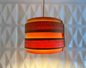 May include: A hanging pendant light fixture with a layered design. The lamp features a red and brown woven shade with an orange glow emanating from within. The background is a white wall with a geometric pattern.