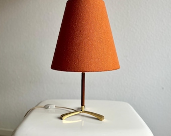 May include: A vintage table lamp featuring an orange fabric lampshade. The lamp has a dark brown stem and a brass-coloured base. The lamp is on a white surface.