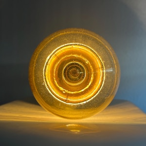 Peut inclure: Gros plan sur une ampoule ronde de couleur ambre. L'intérieur de l'ampoule présente une série de cercles concentriques, avec une lueur vive et chaleureuse émanant de l'intérieur. L'ampoule repose sur une surface claire.