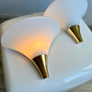 Könnte beinhalten: Zwei weiße Wandleuchten mit goldfarbenem Sockel. Die Leuchten haben eine geschwungene, blütenblattartige Form und strahlen ein warmes Licht aus. Die Leuchten sind auf einer weißen Oberfläche platziert.