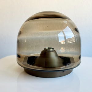 May include: A brown glass decorative object with a rounded top and a metallic base. The glass has a smoky tint, and the base is a darker brown. The object is sitting on a white surface.