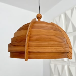 May include: A wooden pendant light fixture with a layered, dome-shaped shade. The light is suspended from a black cord and features a warm, natural wood tone. The design is modern and minimalist, suitable for interior lighting.