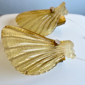 May include: Two vintage wall sconces with a seashell design. The sconces are made of textured, amber-colored glass with a gold finish. Each sconce has a small, round, silver-colored metal accent and a wire for hanging.