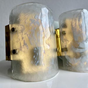 May include: Two vintage wall sconces with a clear, iridescent glass shade and gold-toned metal accents. The sconces have a cylindrical shape and are mounted on a rectangular metal base with visible screws. The glass has a pearlescent finish.