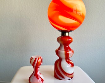 May include: A decorative glass lamp with a spherical orange and white swirled shade. The lamp sits atop a multi-tiered base with a matching color scheme. A smaller, matching vase is next to the lamp. The lamp and vase are on a white surface.