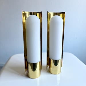 Könnte beinhalten: Zwei moderne Wandleuchten mit goldener Oberfläche und weißen zylindrischen Leuchten. Die Leuchten haben ein schlankes, minimalistisches Design, perfekt, um jedem Raum einen Hauch von Eleganz zu verleihen. Jede Leuchte ist etwa 30 cm hoch.