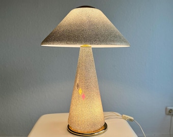 May include: A vintage table lamp with a speckled grey lampshade and a conical base. The base has a textured pattern with small diamond-shaped accents in pink and yellow. The lamp sits on a gold-coloured base.