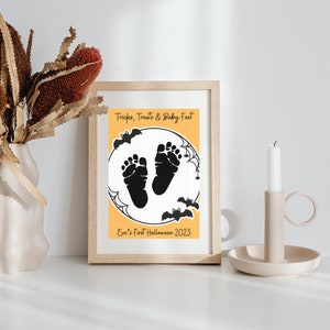 Pode incluir: Uma impress&atilde;o emoldurada com fundo amarelo e silhuetas pretas de p&eacute;s de beb&ecirc;. O texto diz "Tricks, Treats & Baby Feet" e "Eve's First Halloween 2023".