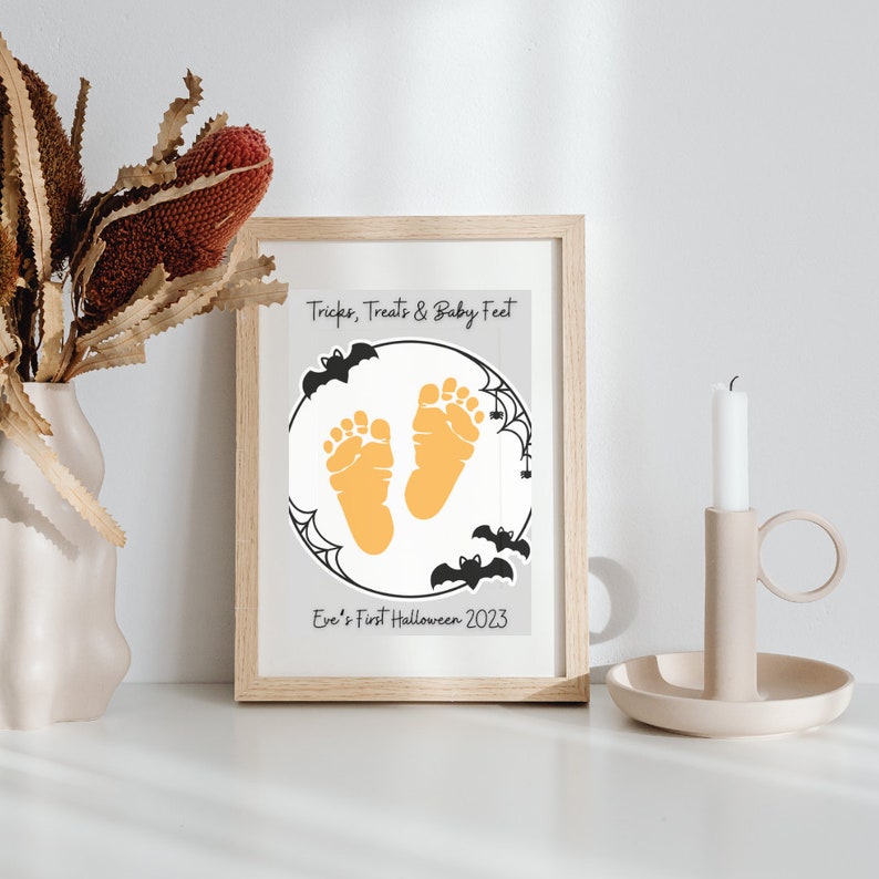 Pode incluir: Uma impress&atilde;o emoldurada com fundo branco e morcegos pretos. A impress&atilde;o apresenta um c&iacute;rculo com duas pegadas de beb&ecirc; laranja dentro. O texto "Tricks, Treats & Baby Feet" est&aacute; no topo da impress&atilde;o e "Eve's First Halloween 2023" est&aacute; na parte inferior.