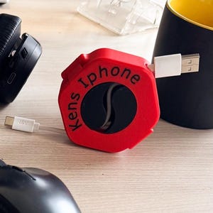 Könnte beinhalten: Roter Kabelorganizer mit dem Text "Kens iPhone" darauf. Der Organizer hat eine sechseckige Form mit einer schwarzen Mitte und einem weißen Kabel, das herauskommt.