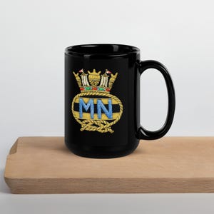May include: Black ceramic mug with a gold and blue embroidered crest featuring the letters "MN" and a crown. The mug is on a wooden surface.