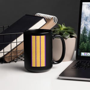 Pode incluir: Uma caneca de café preta com um design vertical de três listras douradas com pequenos pontos, separadas por duas listras roxas. A caneca está em uma superfície branca, com um laptop, livros e uma planta no fundo.