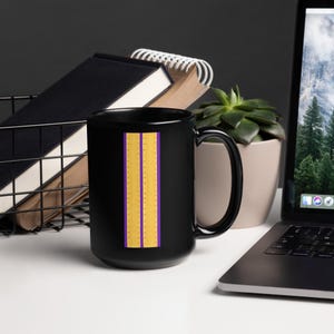 Pode incluir: Uma caneca de café preta com um design vertical de listras douradas e roxas. As listras douradas têm pequenos pontos espaçados uniformemente. A caneca está em uma superfície branca com um laptop, uma planta e material de escritório no fundo.