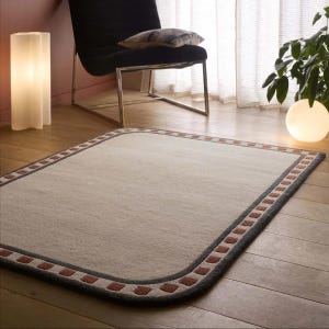 May include: A rectangular rug with rounded corners. The rug features a light beige central area, a dark grey border, and a decorative pattern of small, reddish-brown squares along the edge. It is placed on a wooden floor.