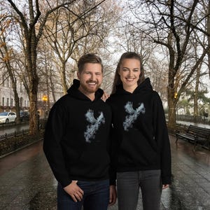 Peut inclure: Deux personnes portant des sweat-shirts à capuche noirs avec un motif blanc d'un oiseau en vol. Les sweat-shirts ont une capuche à cordon et une poche kangourou.