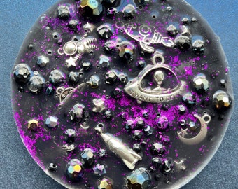 May include: A black wax melt with a space theme. It features silver charms of a rocket, a UFO, a crescent moon, and stars. The wax melt also has purple glitter and black beads.