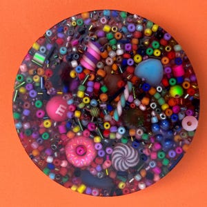 May include: A round resin coaster featuring a vibrant mix of beads, sweets, and small decorative elements. The design includes a pink donut, a pink M&M, and a swirled candy. The background is a bright orange.
