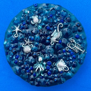 May include: A circular decorative item with a blue resin base, featuring a variety of blue beads and small silver charms. The charms depict sea creatures such as a turtle, seahorse, starfish, and octopus. The background is a solid blue.