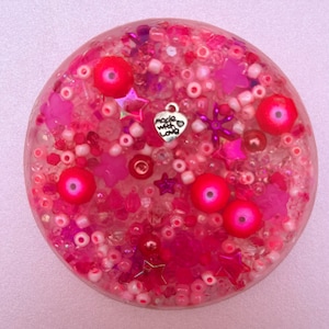 May include: A round, clear resin coaster filled with pink, red, and white beads, stars, and other small decorative elements. A silver charm with the text "Made with Love" hangs from the centre of the coaster.
