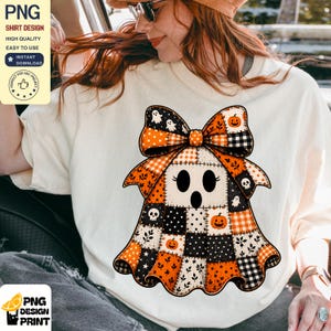 May include: A cream-colored t-shirt featuring a large graphic of a ghost shaped like a patchwork quilt. The ghost has a large bow on top and is decorated with orange and black patterns including pumpkins, skulls, and small ghosts.