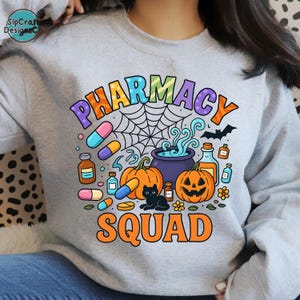 May include: A light gray sweatshirt with a colorful graphic design. The design features the words "PHARMACY SQUAD" in orange, purple, and blue, along with Halloween-themed illustrations of pumpkins, pills, and a black cat.
