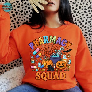 May include: Orange sweatshirt with a colorful "PHARMACY SQUAD" graphic. The design includes pills, pumpkins, a black cat, a cauldron, and a spider web, perfect for Halloween. The sweatshirt is a crew neck.