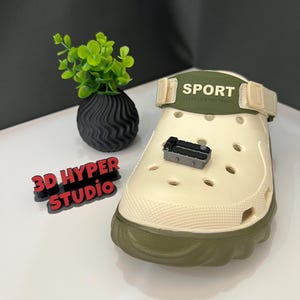Puede incluir: Un zueco beige y verde oliva con la palabra "SPORT" impresa en la correa. Un pequeño objeto negro está encima del zueco. Un pequeño jarrón negro con una planta verde y el texto "3D HYPER STUDIO" también están en la imagen.